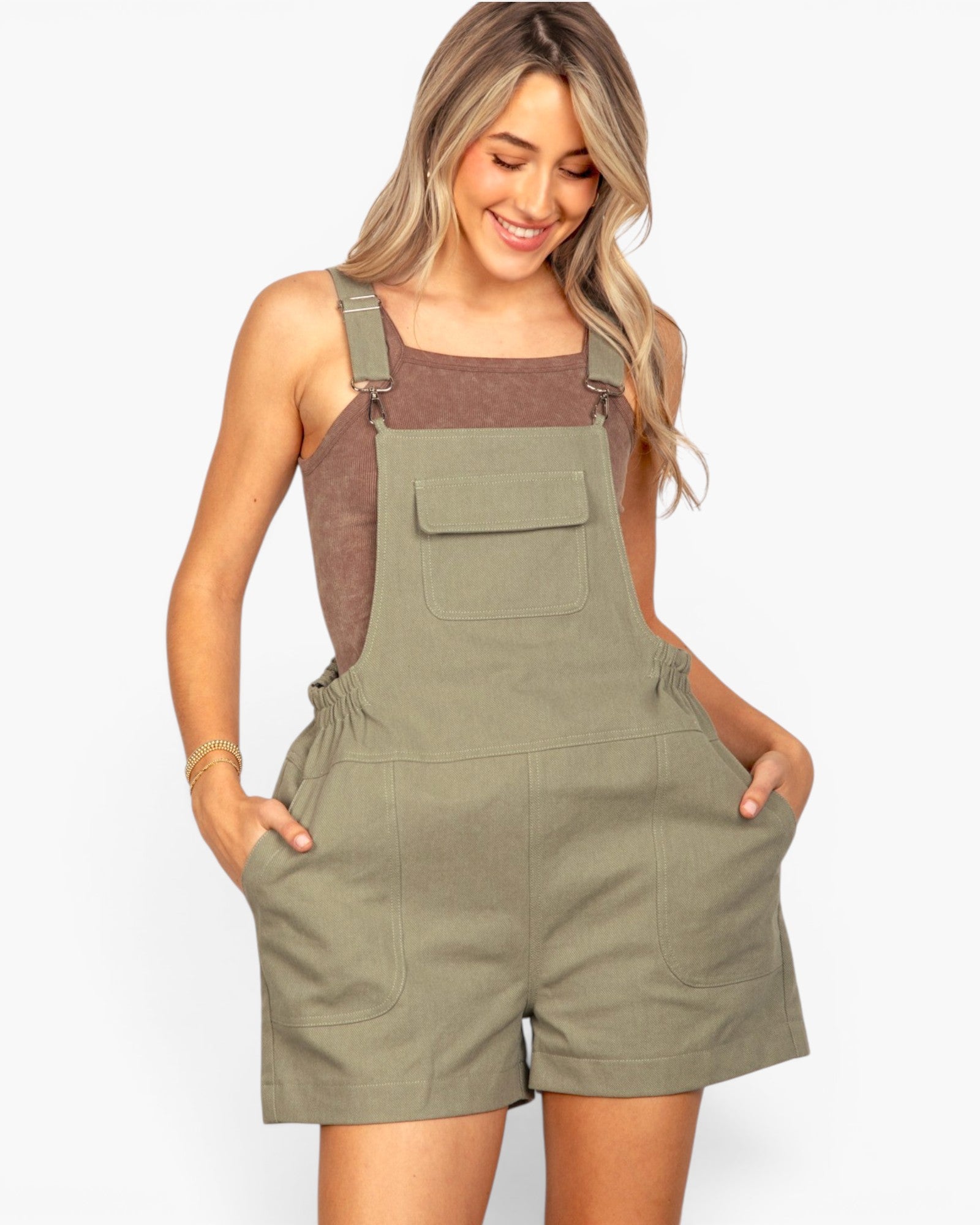 A woman modeling olive green utility-style shorts with shoulder straps and a front flap pocket, worn over a brown tank top against a light gray studio background.