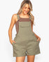 A woman modeling olive green utility-style shorts with shoulder straps and a front flap pocket, worn over a brown tank top against a light gray studio background.