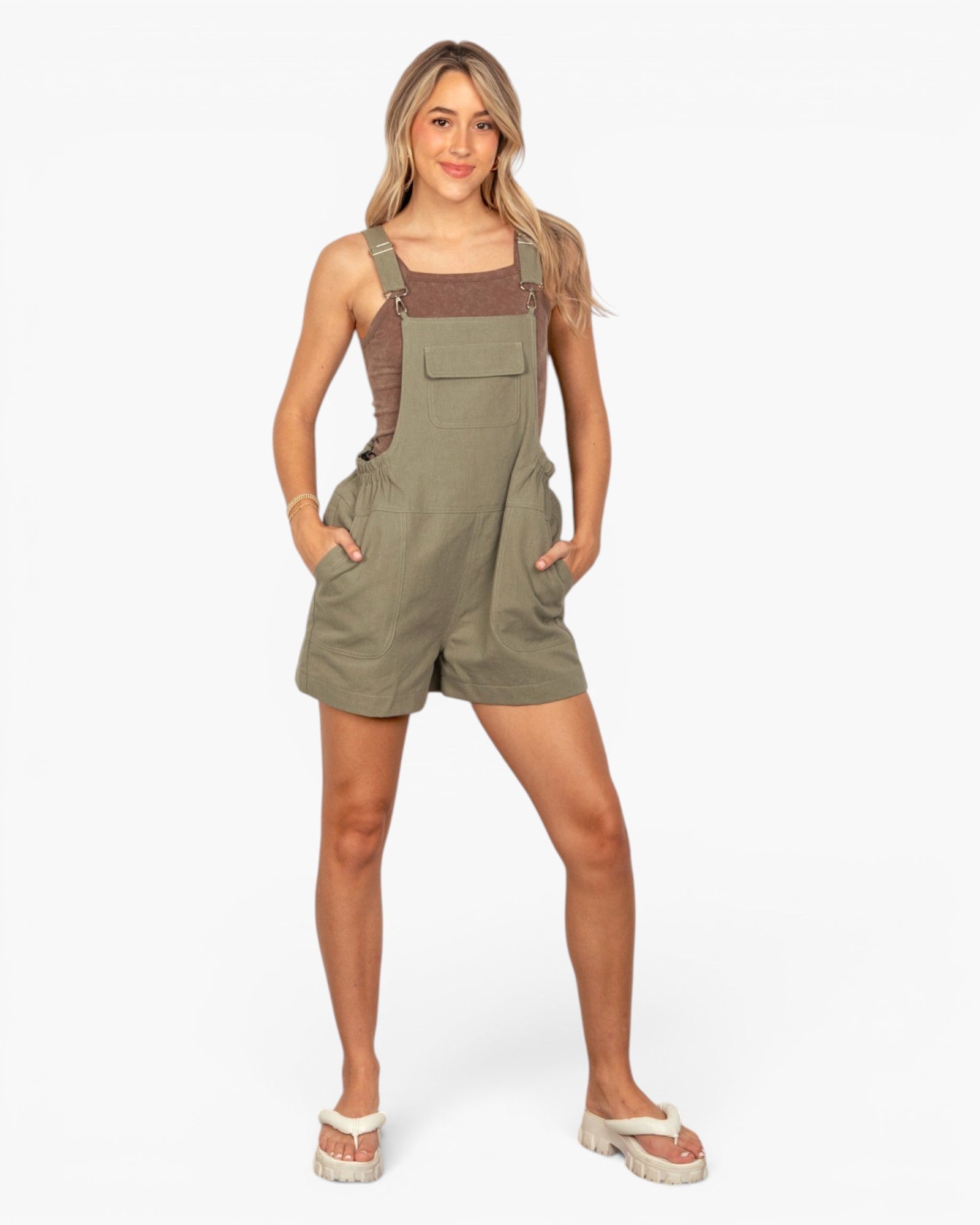 A woman modeling a pair of olive green bib shorts with a brown ribbed tank top underneath, standing against a plain white studio background.