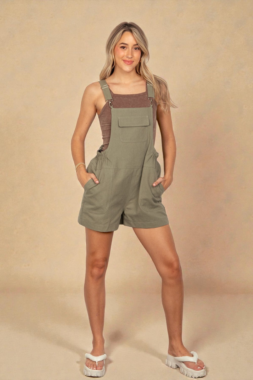 Woman wearing a green overalls with brown straps against a beige background