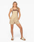 A woman modeling a beige short overall jumpsuit with a brown tank top underneath and beige slide sandals, set against a plain white studio background.