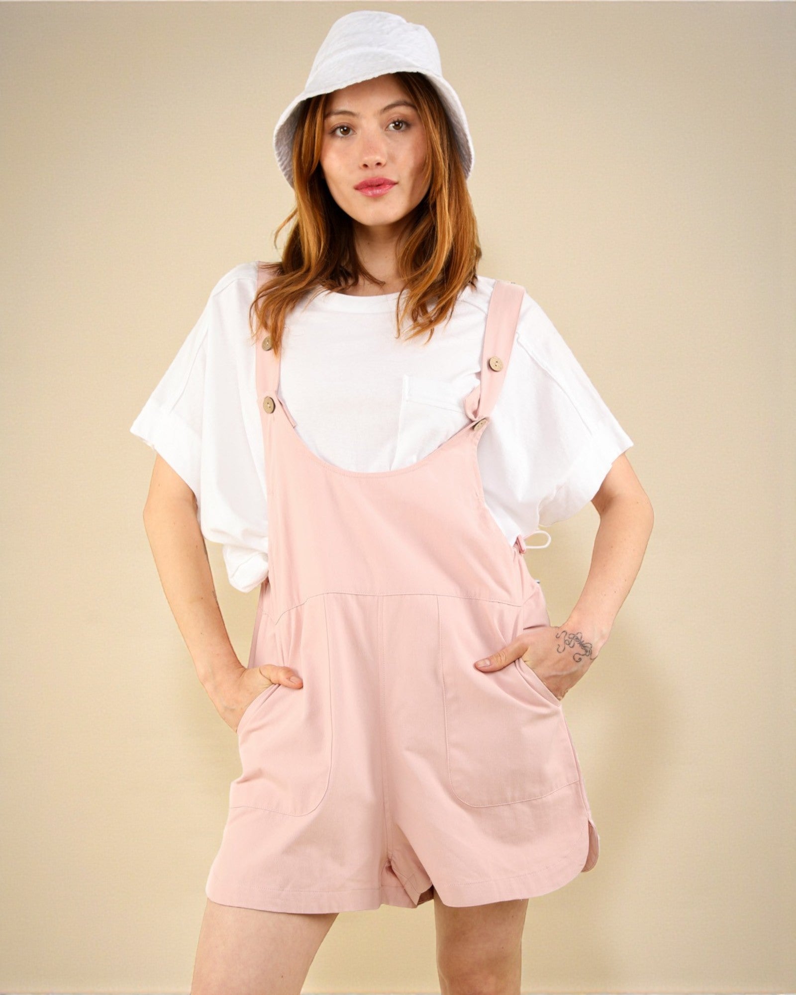 Person wearing a white shirt, pink overalls, and a white bucket hat on a beige background