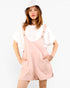 A woman modeling light pink overall shorts with a white t-shirt and a white bucket hat, standing against a plain white background.