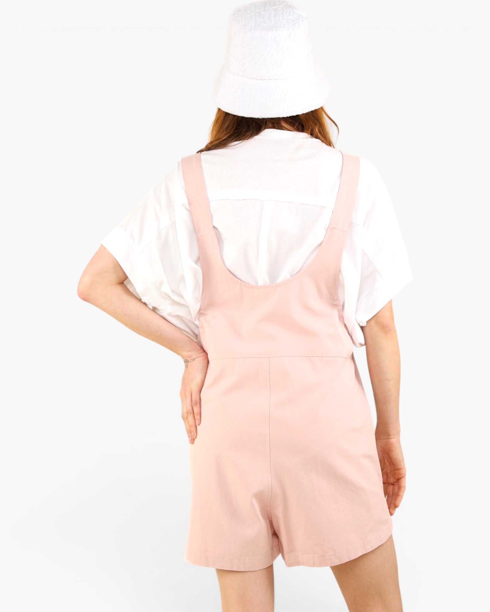 A model shown from the back wearing a light pink overall romper over a white short-sleeve shirt, with a white bucket hat, against a plain white background.