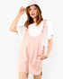 A woman modeling a light pink overall romper over a white t-shirt, wearing a white bucket hat, against a plain white background.