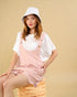 Woman wearing a pink pinafore dress over a white shirt with a white bucket hat on a beige background