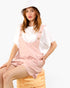 A woman wearing a light pink overall romper over a white t-shirt and a white bucket hat, sitting on a wooden barrel stool against a plain white background.