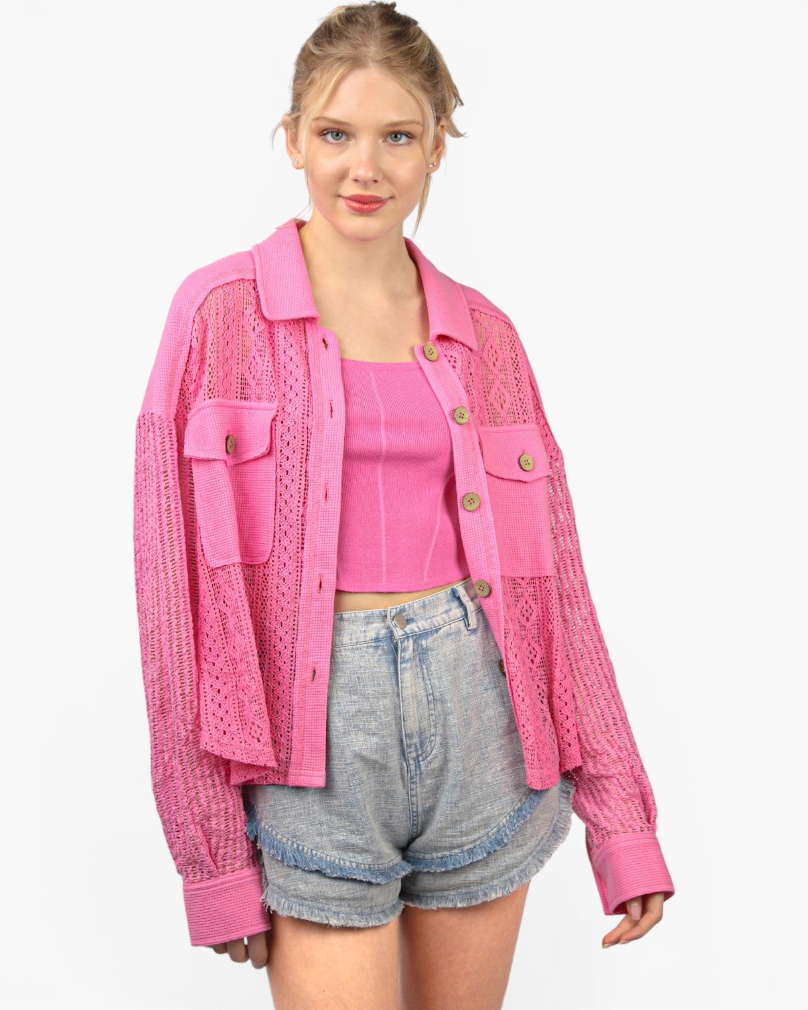 A woman modeling a bright pink open-front knit cardigan with button details and chest pockets, worn over a matching pink crop top and light-wash denim shorts, against a plain white background.