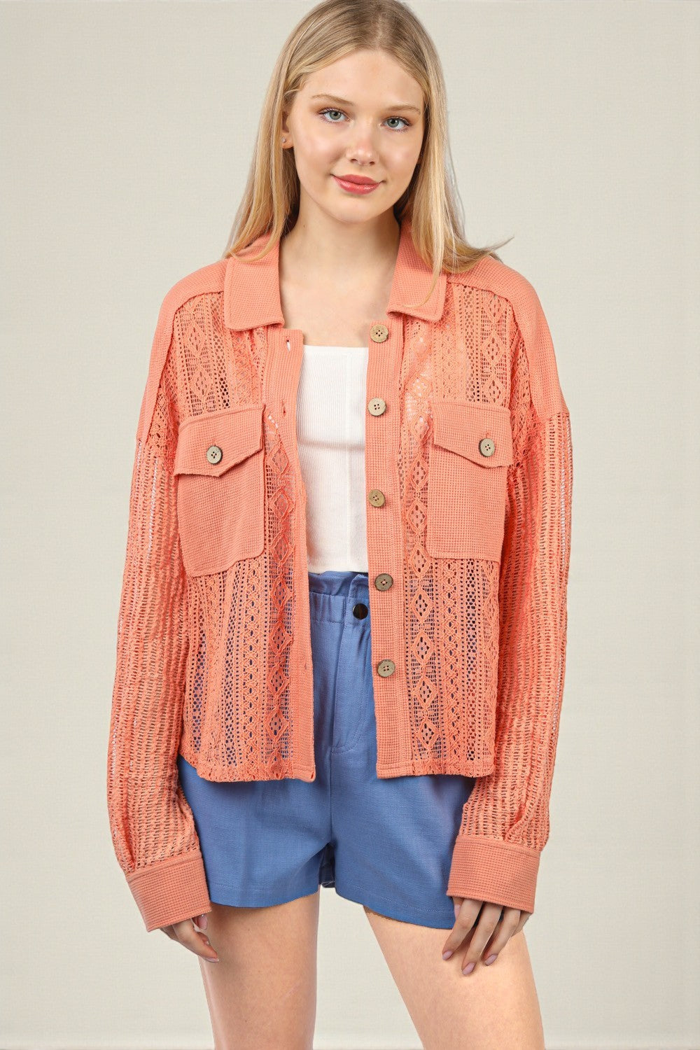 Woman wearing a coral lace jacket over a white top and blue shorts on a beige background
