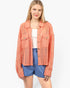 A woman modeling a coral pink open-front jacket with textured knit and woven panels, worn over a white top and blue shorts against a light gray studio background.