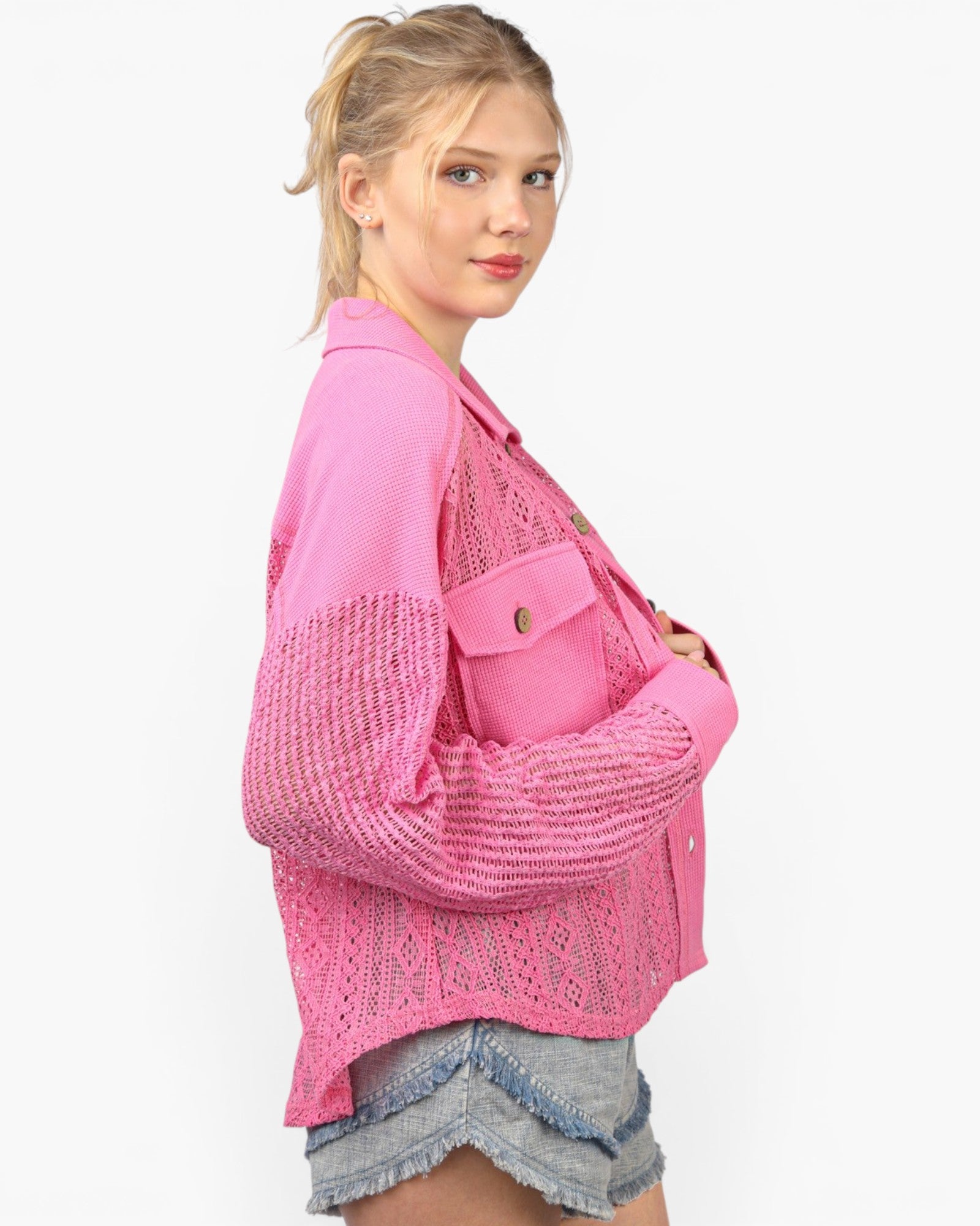 A woman wearing a bright pink knit cardigan with textured knit sleeves and a smooth knit body, paired with denim shorts, against a plain white background.