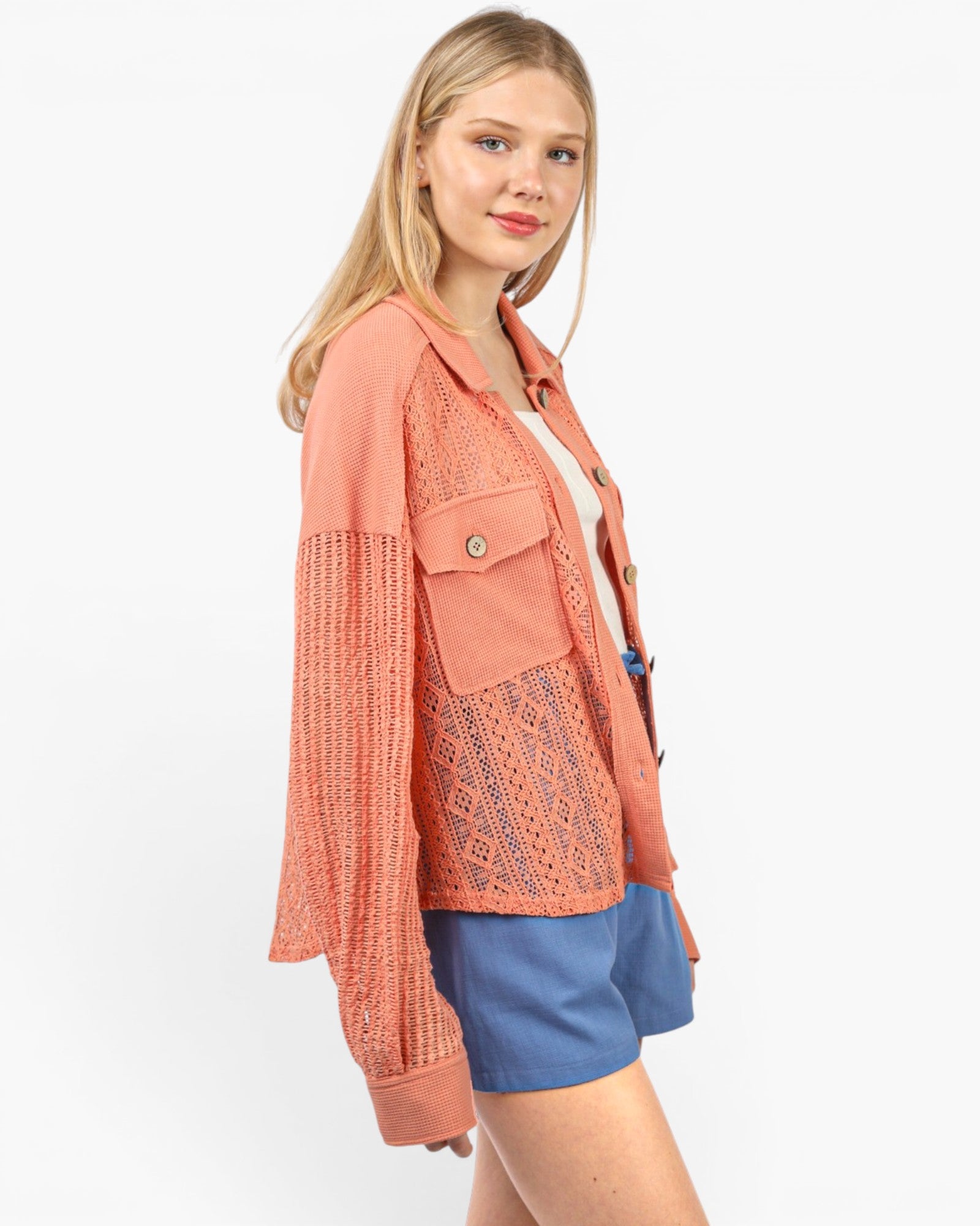 A woman modeling a coral orange textured knit jacket with long sleeves, a button-up front, and a chest pocket, paired with blue shorts against a plain white background.