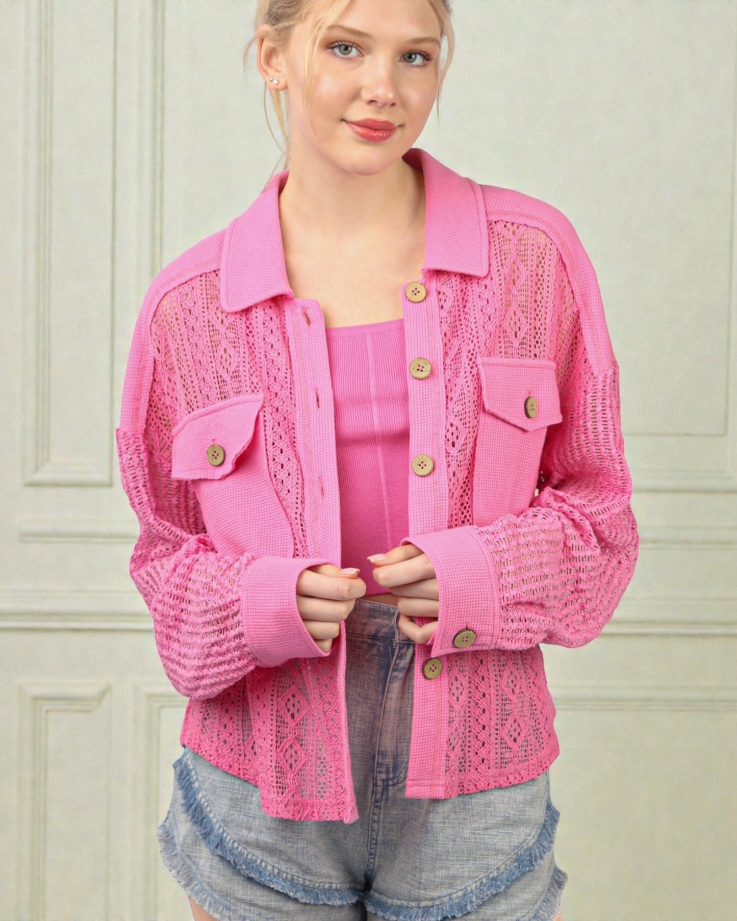 Woman wearing a pink lace jacket with gold buttons against a light-colored wall.