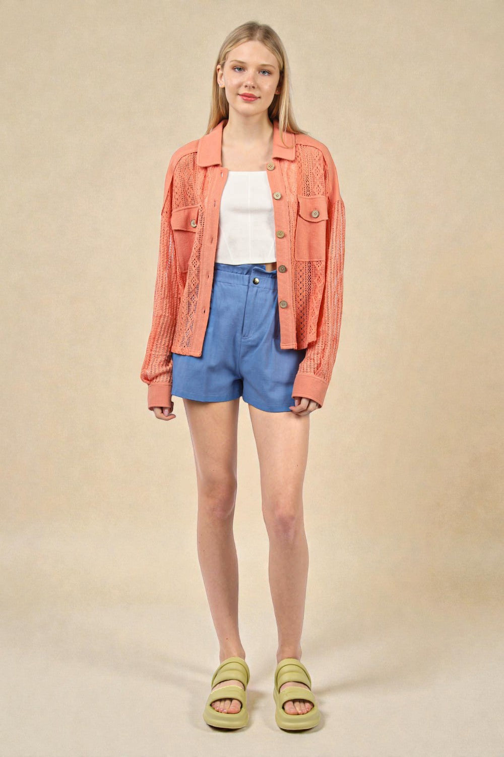 Woman wearing a coral jacket, white top, blue shorts, and green sandals on a beige background
