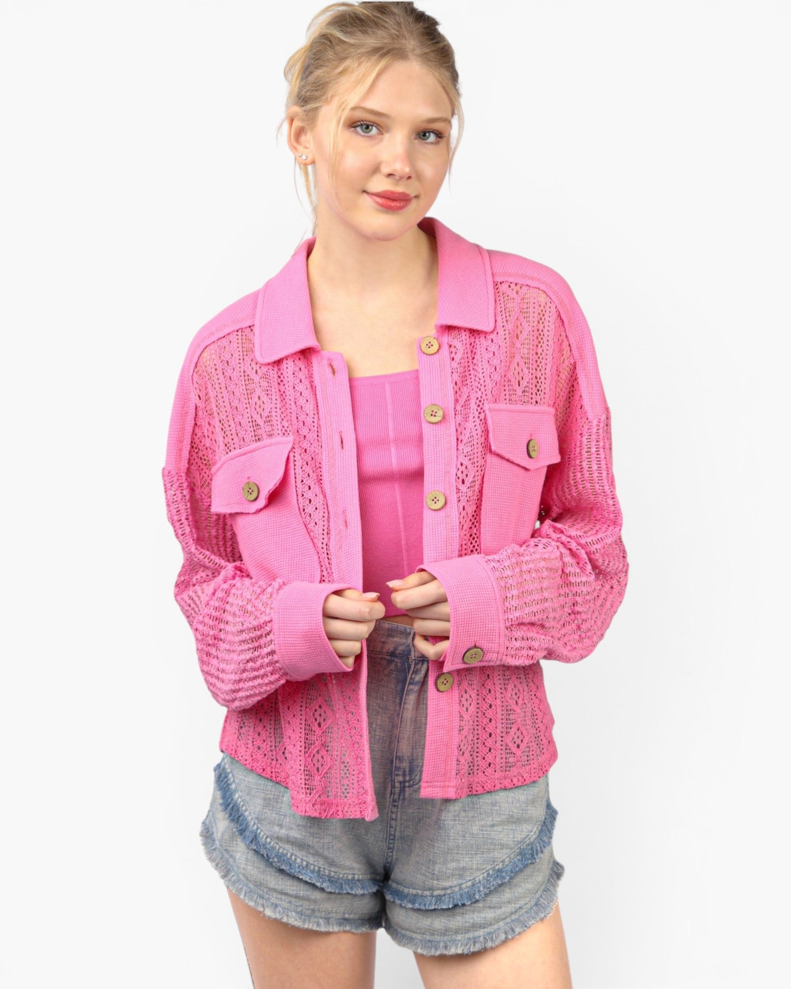 A woman modeling a bright pink open-front knit cardigan with a collar, chest flap pockets, and gold-tone buttons, worn over a matching pink top and denim shorts against a light gray studio background.