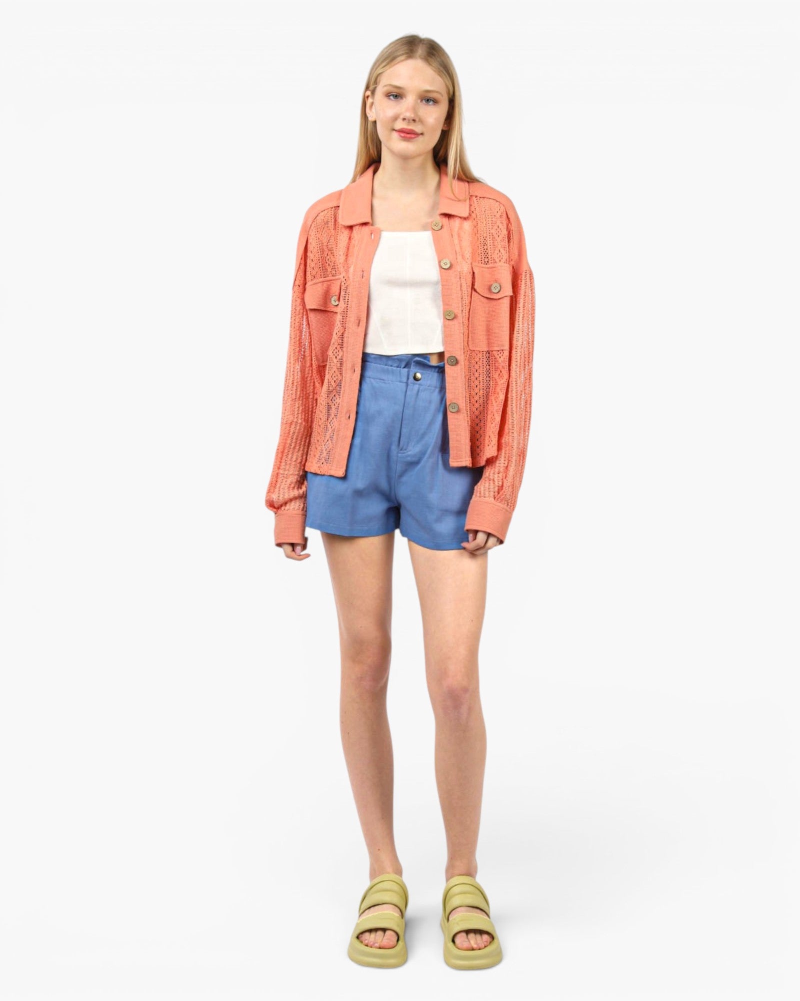 A young woman modeling a coral pink textured jacket over a white tank top, paired with blue denim shorts and olive green slide sandals, against a plain white studio background.