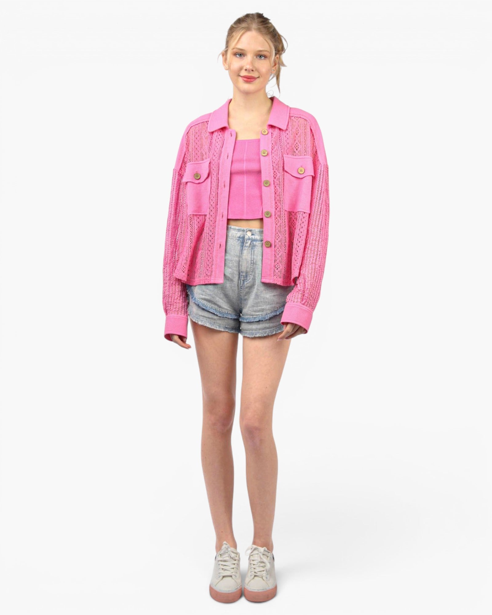 A woman modeling a bright pink textured knit jacket over a matching pink crop top, paired with denim shorts and white sneakers, against a plain white studio background.