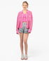 A woman modeling a bright pink textured knit jacket over a matching pink crop top, paired with denim shorts and white sneakers, against a plain white studio background.