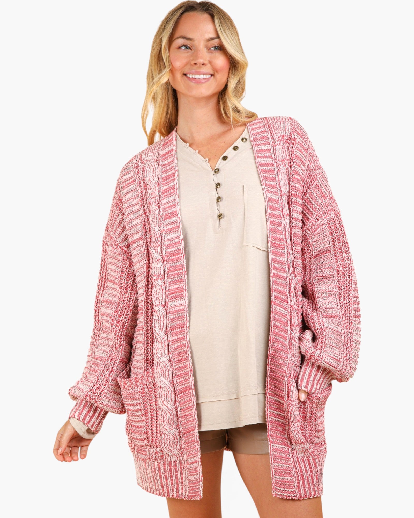 A woman modeling a pink and white textured knit cardigan with a V-neck, button details, and front pockets, worn over a beige henley top against a light gray background.