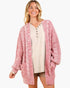 A woman modeling a pink and white textured knit cardigan with a V-neck, button details, and front pockets, worn over a beige henley top against a light gray background.