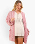 A woman wearing a pink and white textured knit cardigan over a beige henley top and shorts, photographed against a clean white studio background.
