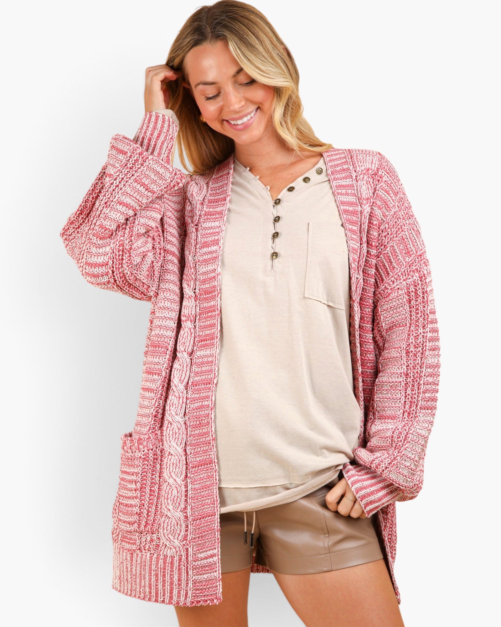 A woman modeling a pink and white marled knit cardigan with a buttoned henley shirt and metallic shorts against a plain white background.
