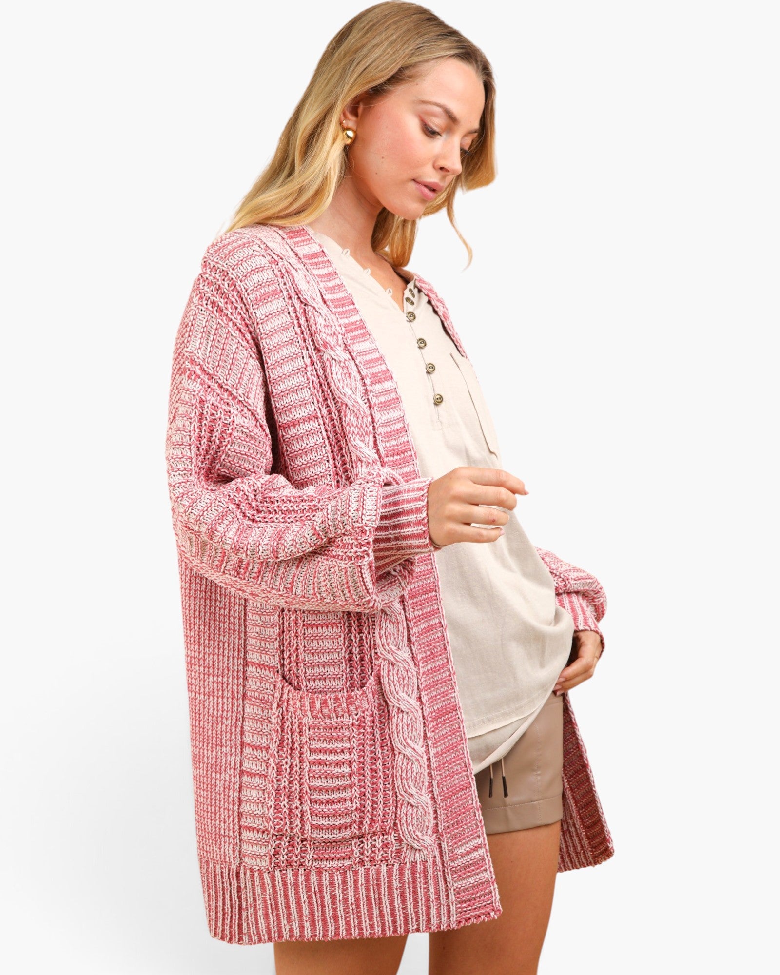 A woman modeling a pink and white textured cable-knit cardigan over a cream button-up top and tan shorts, set against a plain white studio background.