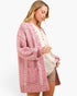A woman modeling a pink and white textured cable-knit cardigan over a cream button-up top and tan shorts, set against a plain white studio background.