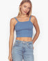 A woman modeling a blue ribbed knit crop top with thin shoulder straps and a square neckline, paired with light-wash blue jeans against a plain white studio background.