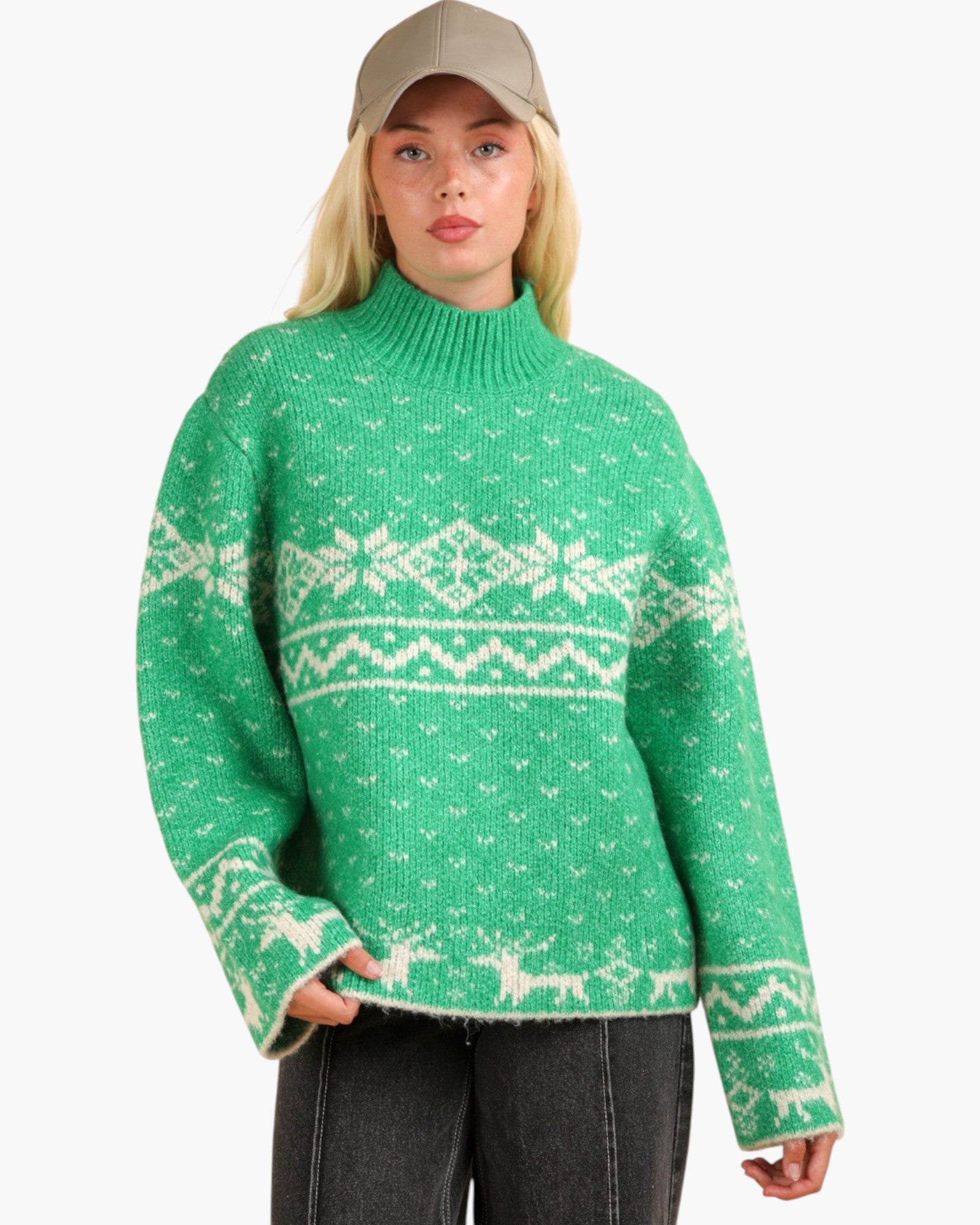 A woman wearing a green knit sweater with a white Nordic fair isle pattern, paired with a beige baseball cap and dark gray jeans, against a plain white background.