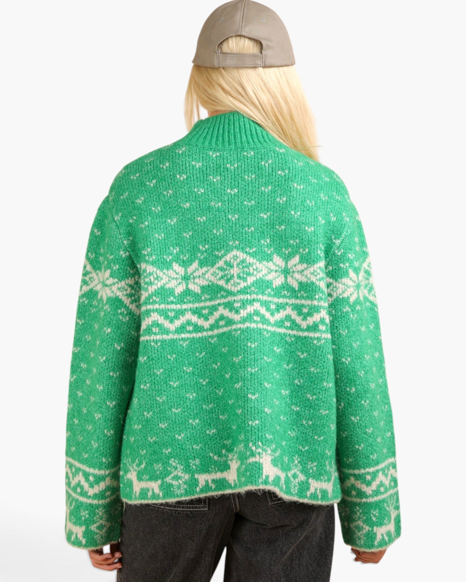 A person shown from the back wearing a green knit cardigan with a white Fair Isle pattern featuring snowflakes, geometric bands, and reindeer silhouettes. The person has blonde hair, wearing a beige baseball cap backwards and dark gray jeans, against a plain white studio background.