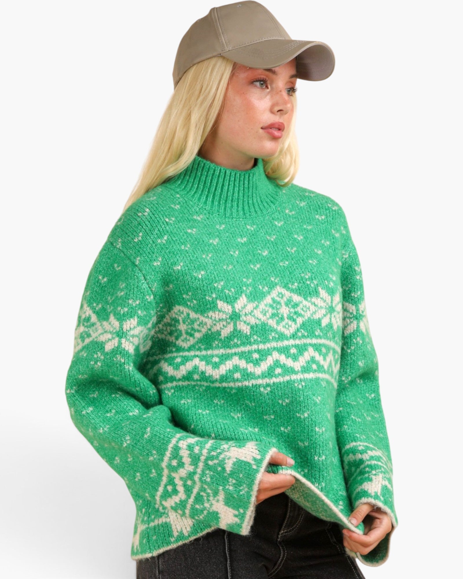 A woman wearing a green knit sweater with a cream Nordic pattern, paired with a beige baseball cap and dark jeans, shown against a plain white background.