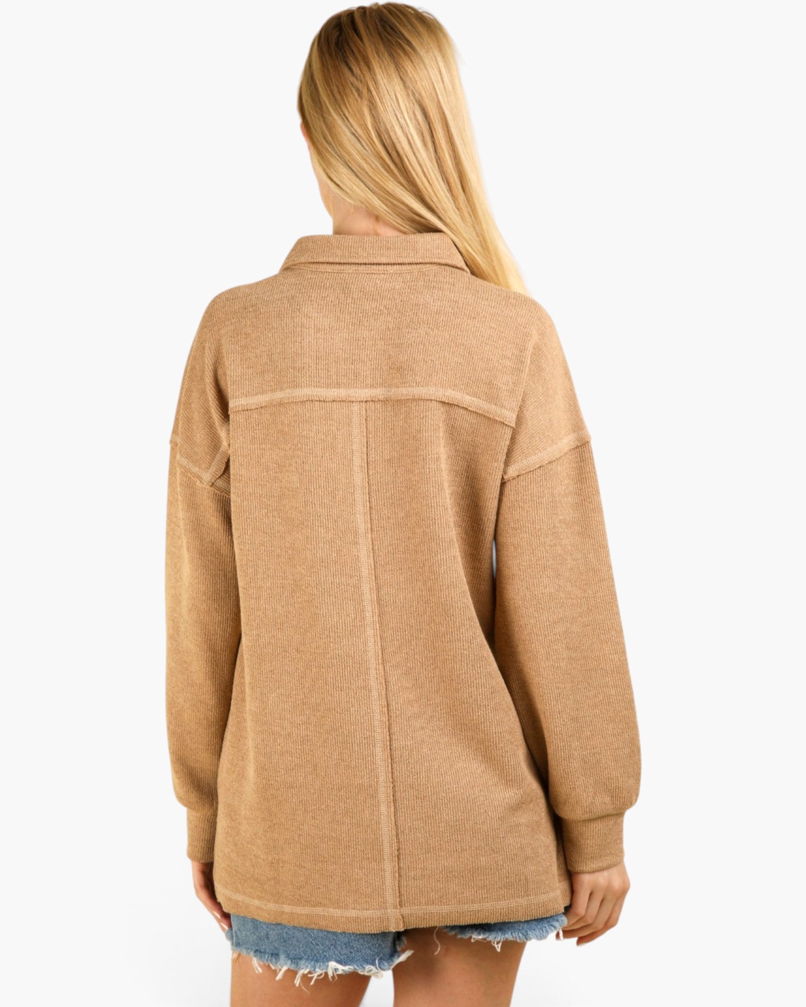 A woman shown from the back wearing a camel-colored, oversized long-sleeve button-up shirt jacket with raw-edge detailing, paired with frayed denim shorts, against a plain white background.