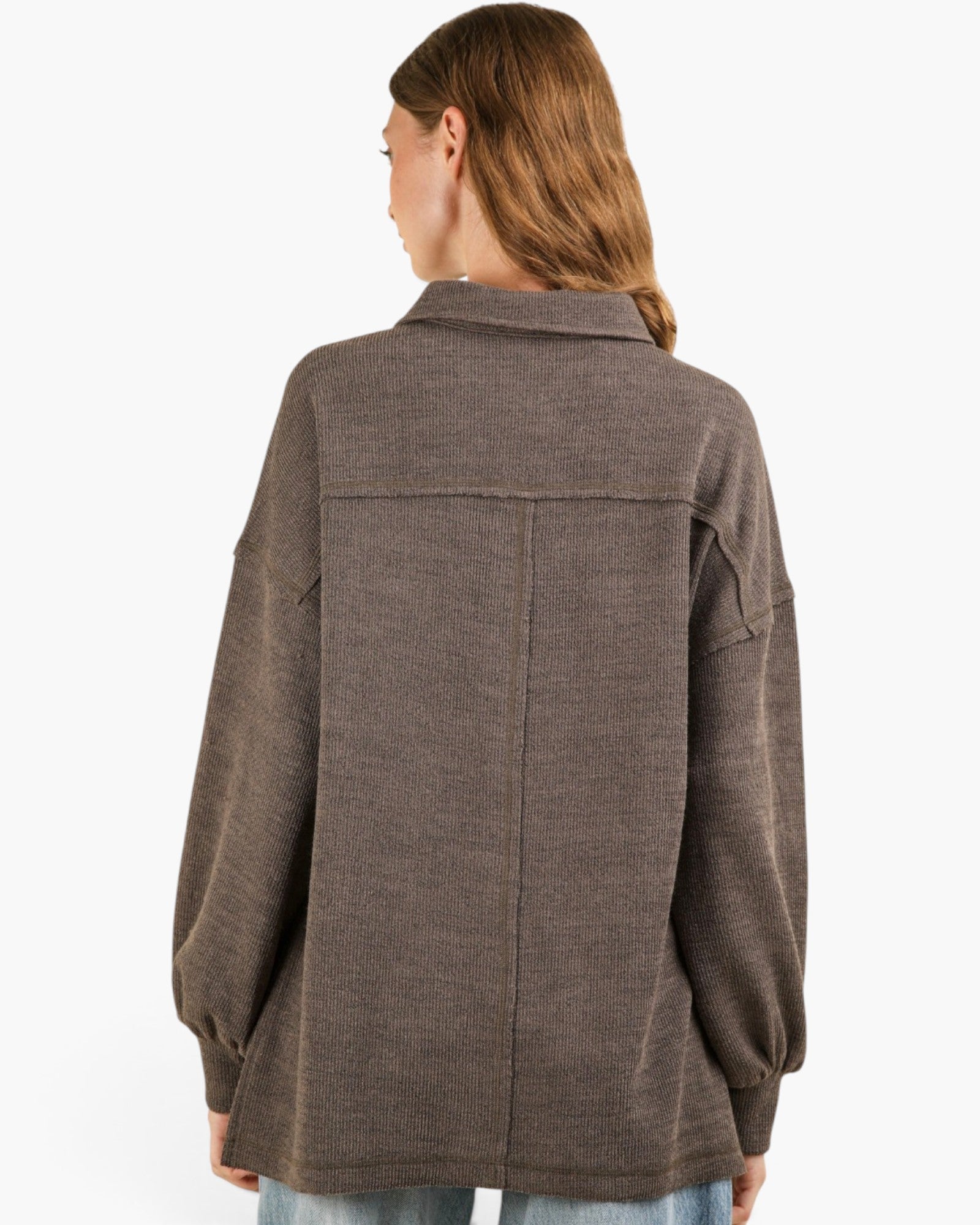 A woman viewed from the back wearing an oversized brown knit button-up shirt jacket with a collar and long sleeves, paired with light blue jeans against a plain white background.