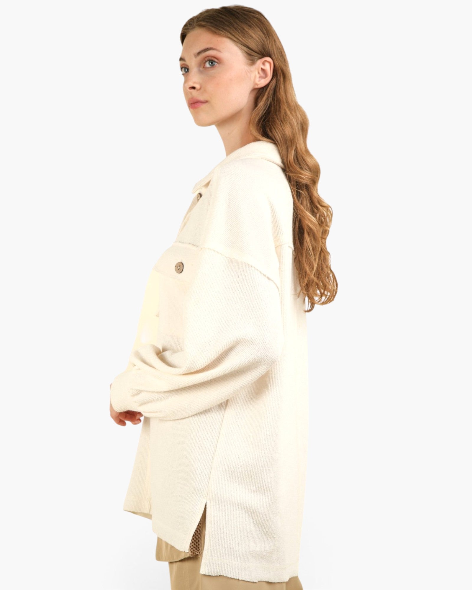 A woman with long wavy light brown hair is shown in profile wearing an oversized cream-colored knit cardigan with a single visible brown button. The background is a clean, solid white studio backdrop.