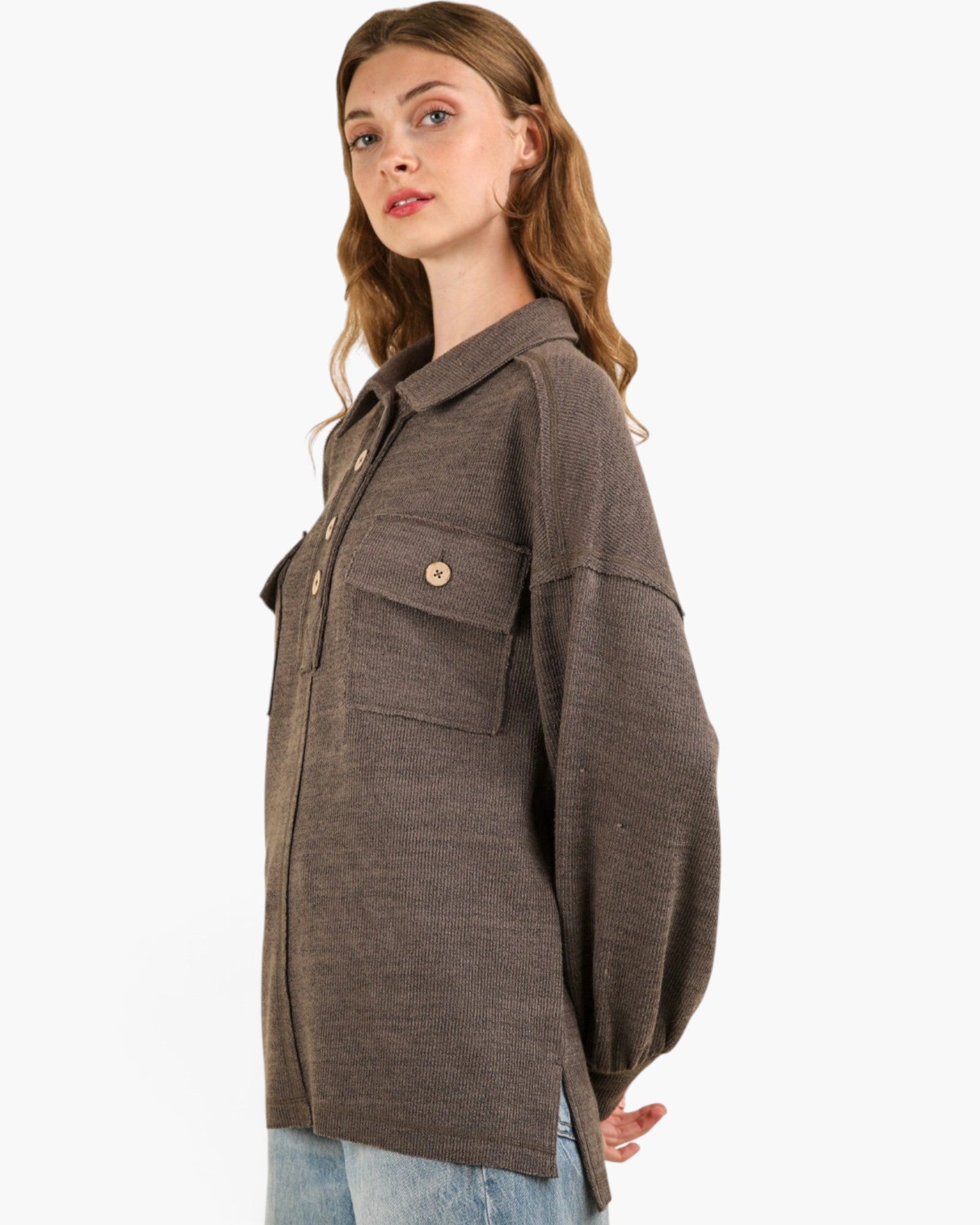 A woman wearing an oversized brown knit button-up shirt jacket with a chest pocket and button, paired with light blue jeans, photographed against a plain white background.
