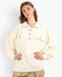 A woman modeling a cream-colored, long-sleeve button-up shirt with two chest flap pockets and brown buttons, paired with beige pants, against a light gray studio background.