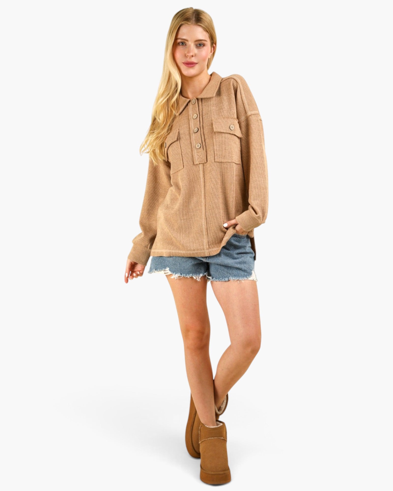 A woman modeling a beige oversized knit button-front pullover with a collar and chest pockets, paired with denim shorts and brown boots, against a light gray studio background.