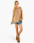 A woman modeling a beige oversized knit button-front pullover with a collar and chest pockets, paired with denim shorts and brown boots, against a light gray studio background.