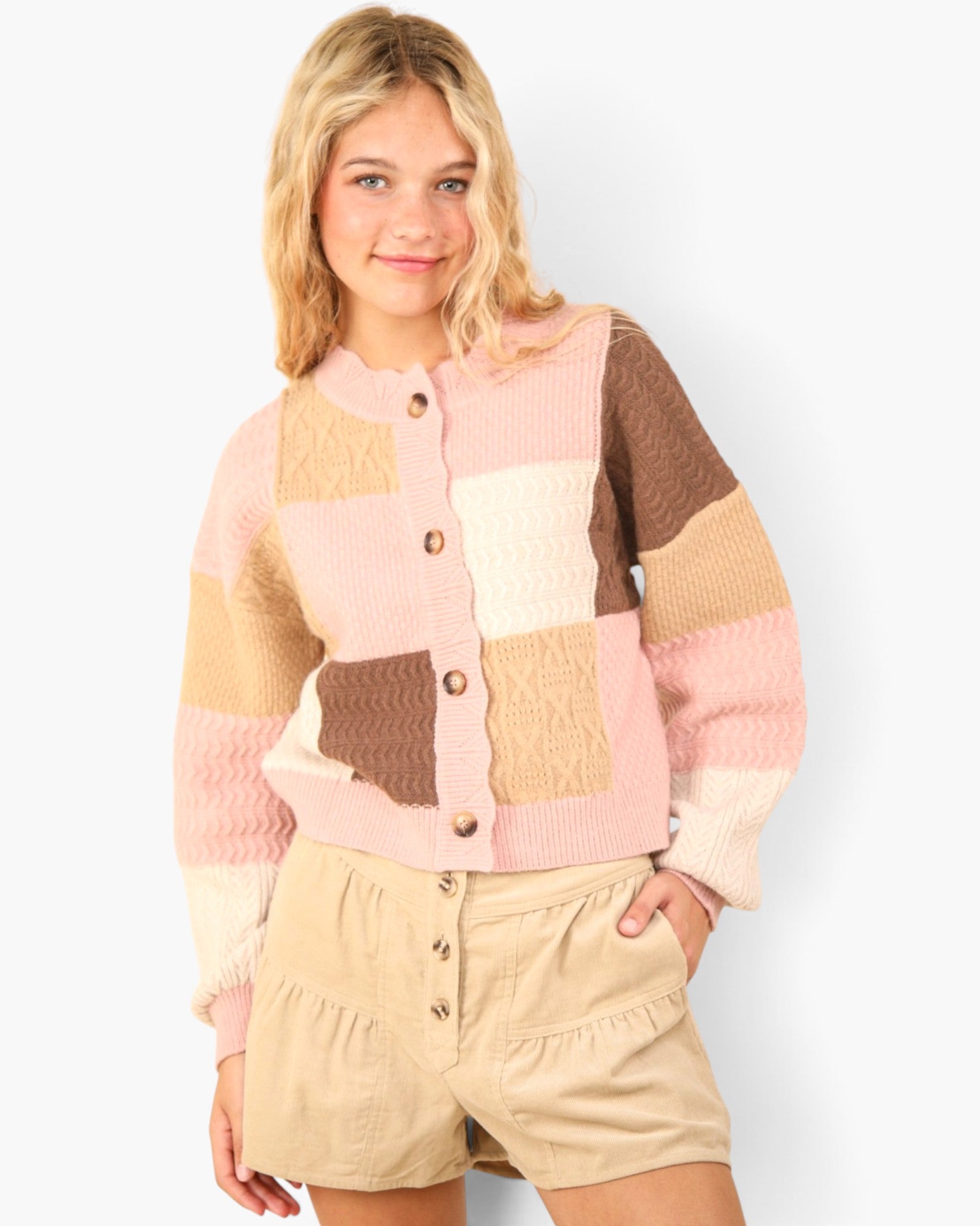 A young woman modeling a pastel patchwork knit cardigan with brown buttons, paired with beige shorts, against a light gray studio background.