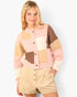 A young woman modeling a pastel patchwork knit cardigan with brown buttons, paired with beige shorts, against a light gray studio background.
