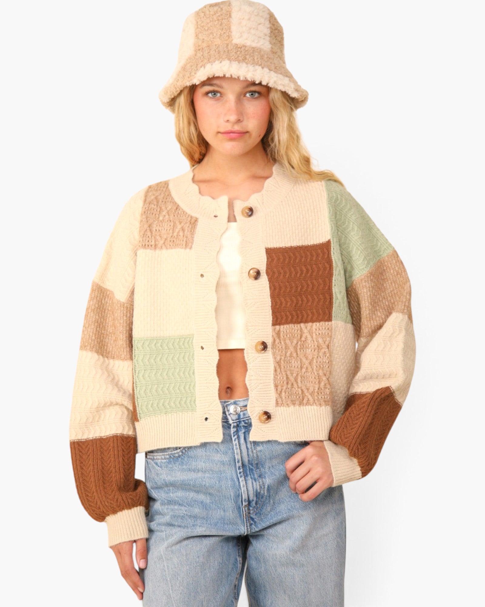 A woman wearing a cream, brown, and sage green patchwork knit cardigan with a matching knit bucket hat and light blue jeans, photographed against a solid white background.
