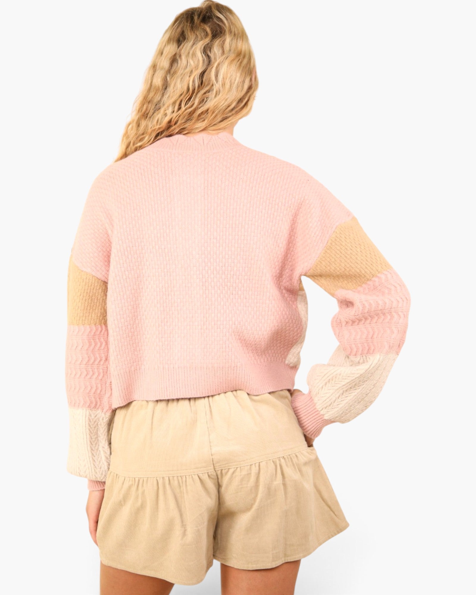A woman shown from the back wearing a pastel pink knit sweater with beige and cream color-blocked sleeves, paired with beige shorts, against a plain white background.