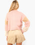 A woman shown from the back wearing a pastel pink knit sweater with beige and cream color-blocked sleeves, paired with beige shorts, against a plain white background.