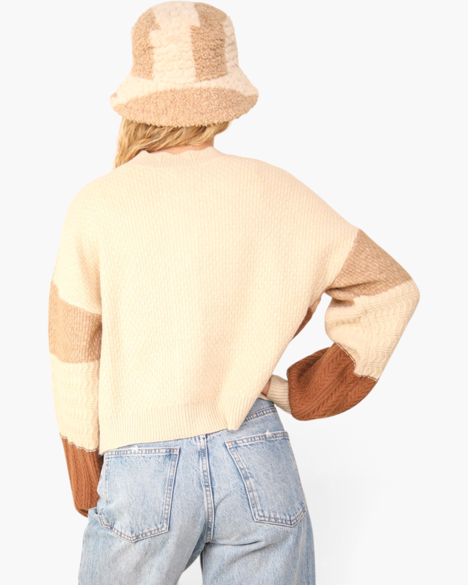 A person shown from the back wearing a cream and tan knit sweater with brown elbow patches, a matching beige bucket hat, and light blue jeans against a plain white background.