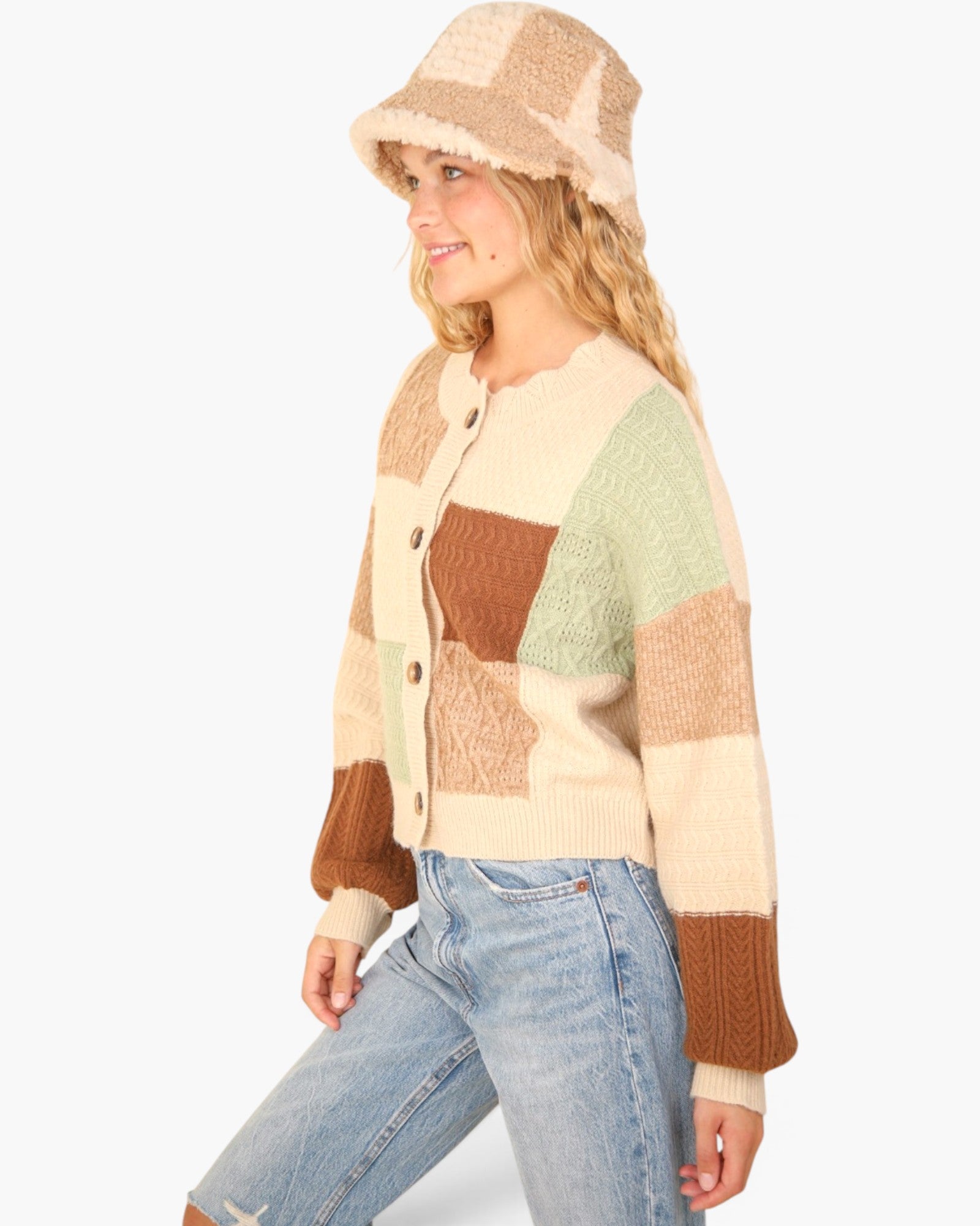 A woman wearing a cream, brown, and sage green patchwork knit cardigan, a matching beige knit bucket hat, and light blue jeans, photographed against a solid white background.