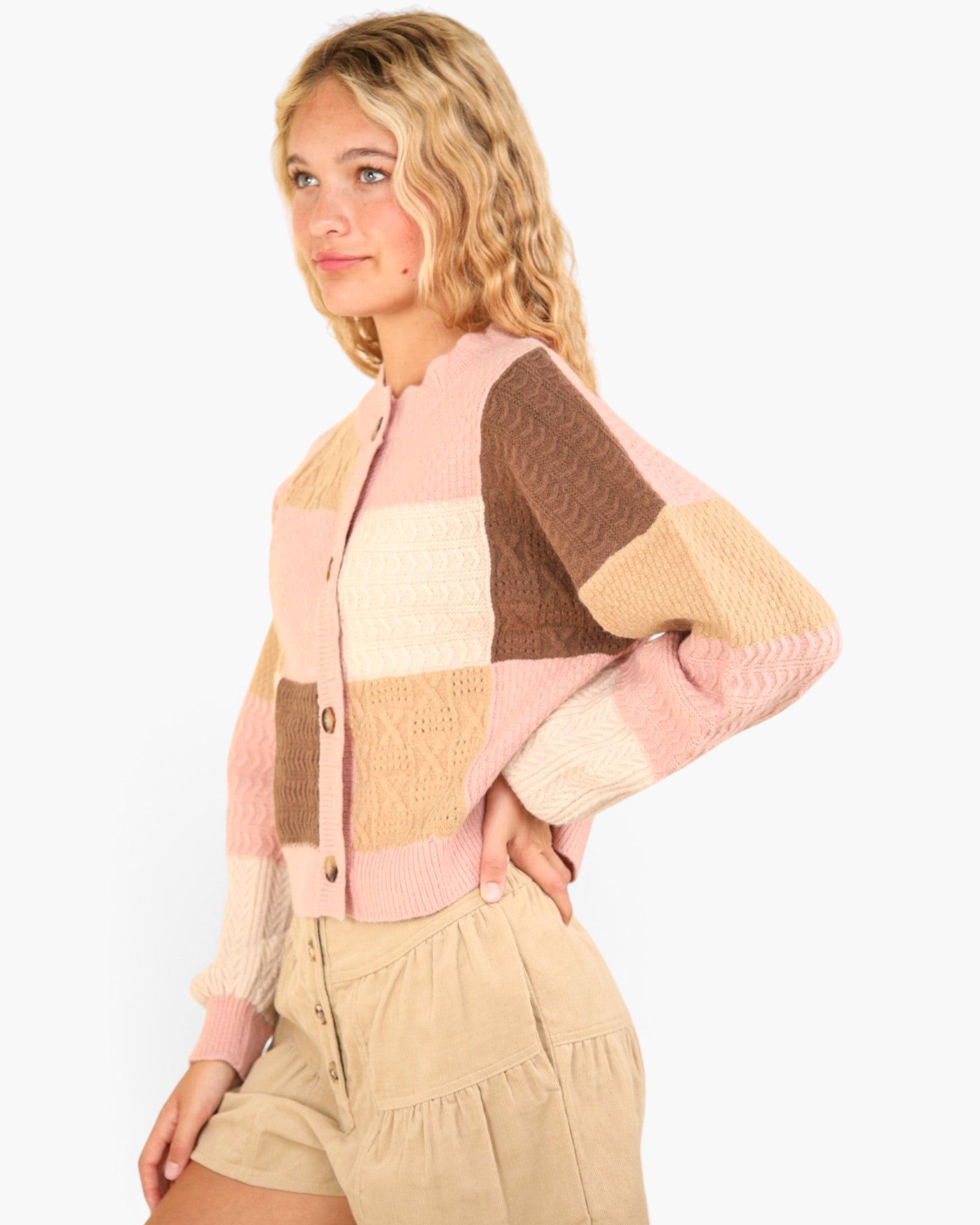 A woman wearing a pastel patchwork knit cardigan with pink, beige, and brown blocks, paired with beige shorts, against a plain light gray background.