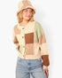 A woman wearing a cream, tan, brown, and sage green patchwork knit cardigan with a matching knit bucket hat and light blue jeans, photographed against a clean white background.