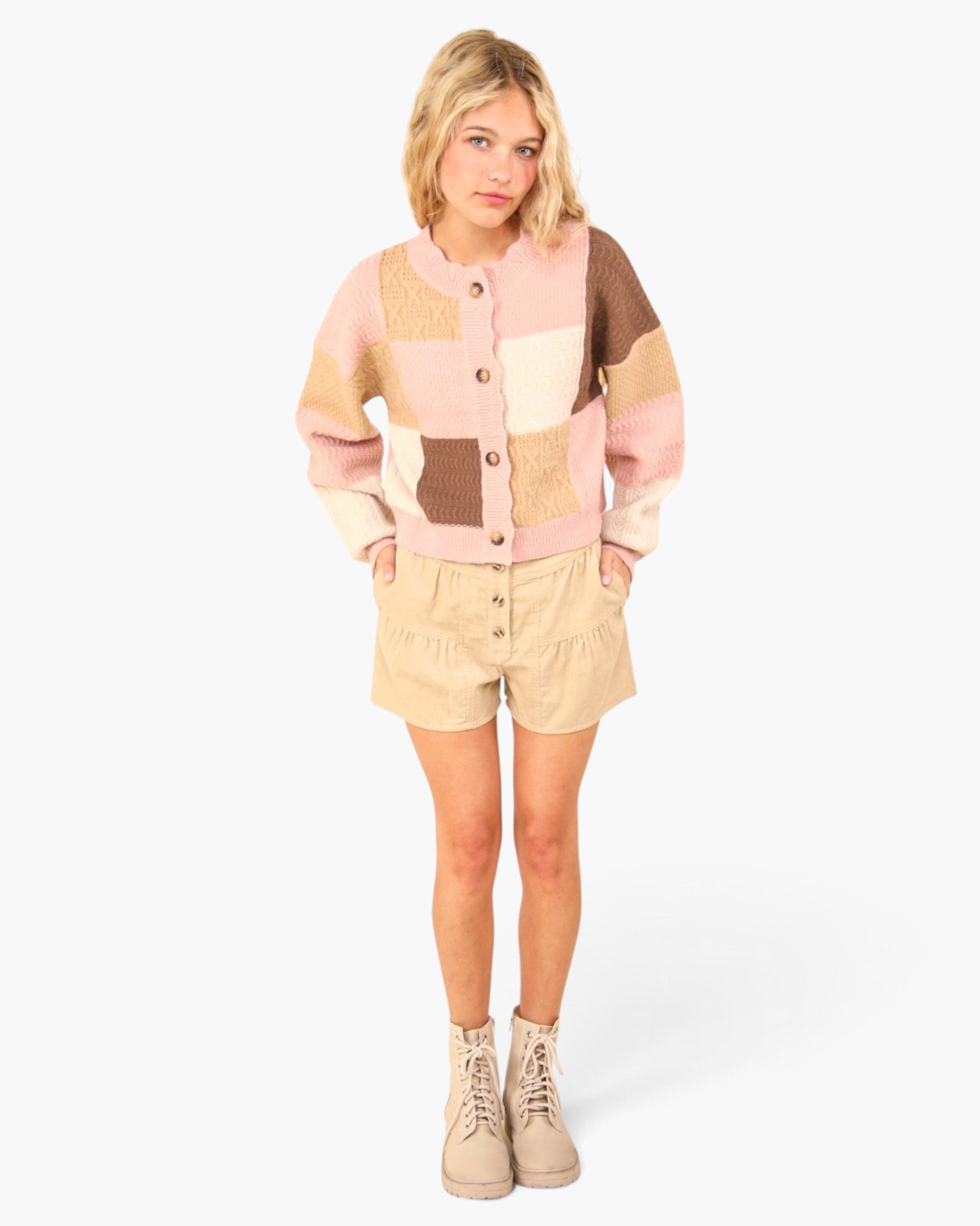 A young girl stands against a light gray studio background wearing a pastel patchwork cardigan with beige shorts and beige lace-up boots. The cardigan features blocks of light pink, cream, and brown.