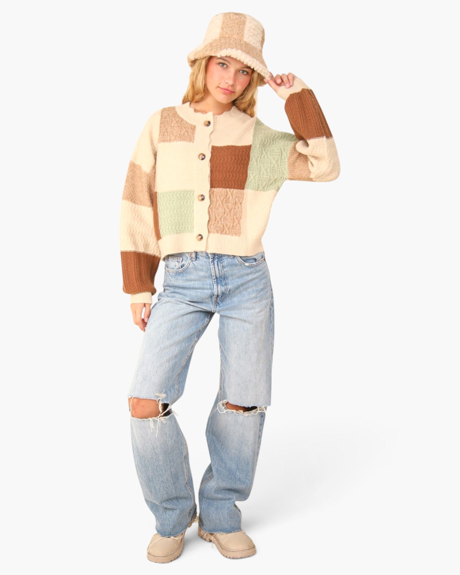 A woman modeling a cream and brown patchwork knit cardigan with a beige fringed bucket hat and distressed light-wash jeans, standing against a plain white studio background.